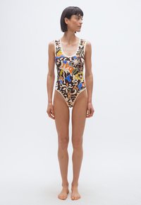 Γυναίκα που στέκεται ξυπόλητη φορώντας ένα καφέ ολόσωμο μαγιό με πολύχρωμα αφηρημένα φυτικά και πουλιών μοτίβα, κοιτώντας πλάγια σε ένα απλό φόντο.