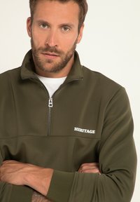 Bluza oliwkowa na zamek z wysokim kołnierzem, białym napisem logo "HERITAGE" oraz ściągaczami na rękawach. Gładka faktura tkaniny.