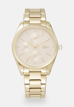 Montre dorée avec un cadran rond, trois sous-cadrans, des accents en diamant et un bracelet en métal poli. La montre présente une lunette texturée et un logo.