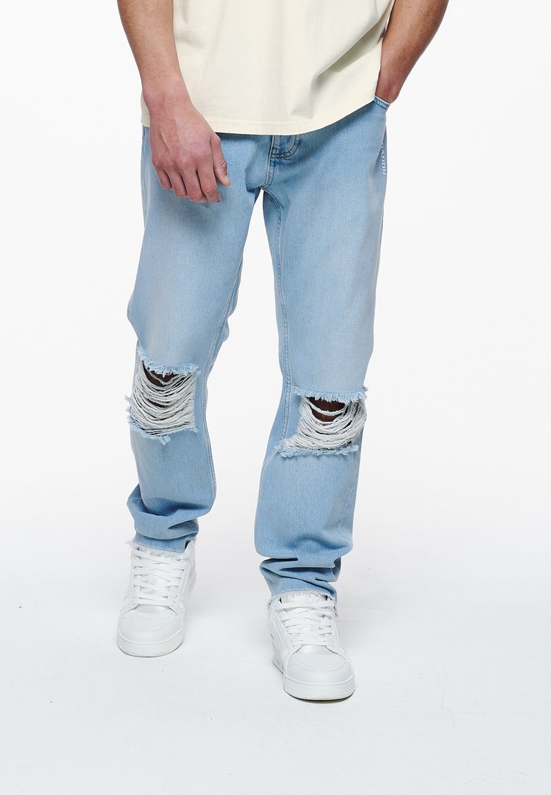 Σαπορές τζιν μπούκλες σε ανοιχτό μπλε χρώμα με σχισμές στα γόνατα, άνετη εφαρμογή, συνδυασμένες με λευκά αθλητικά παπούτσια και κρεμ μπλούζα.