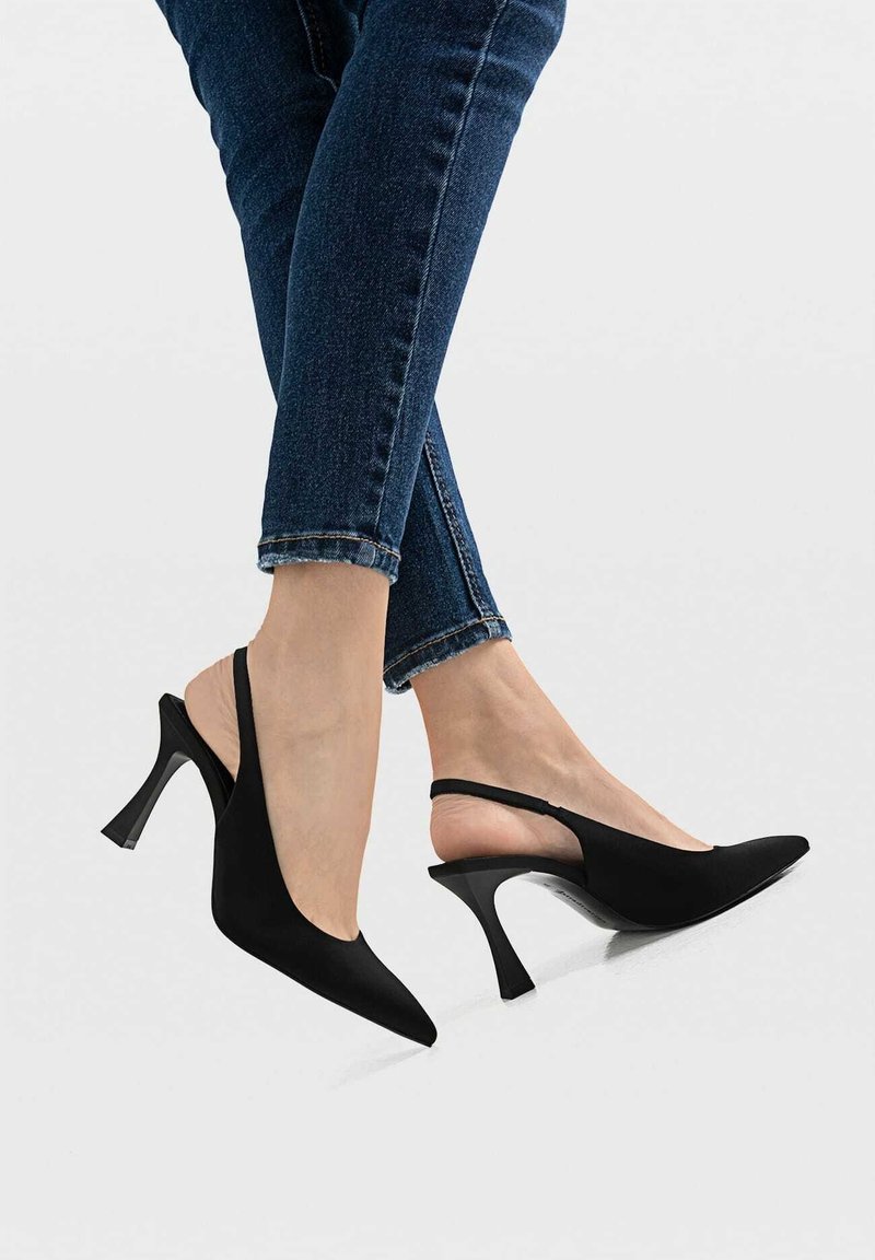 Svarta slingbackklackar med spetsiga tår, texturerad tyg och en dekorativ böjd klack. Bärs med mörkblå jeans med en beskuren fåll.