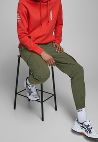 Röd huvtröja med dragsnören, vit text, olivgröna militärbyxor och gråvita sneakers med orange detaljer, sittande på en svart pall.