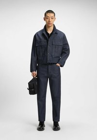 Giacca di jeans e pantaloni in blu scuro, con un design corto, grandi tasche frontali e una texture liscia. Borsa nera con chiusura tenuta in mano.