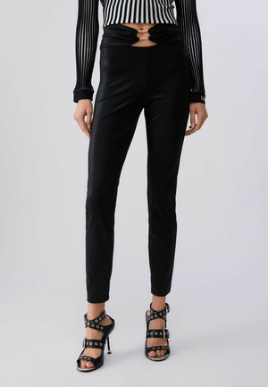 Žena nosiace čierne nohavice s vysokým pásom a úzkym strihom s výrezom a zlatou sponou v páse, v kombinácii s čiernymi sandálmi na podpätku so sponami.