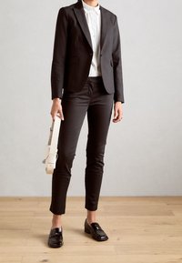 Blazer noir ajusté porté sur une chemise blanche boutonnée, associé à un pantalon noir coupe slim et des mocassins noirs brillants. Tenant un sac blanc.