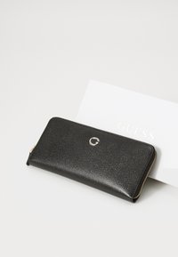 Portefeuille en cuir noir avec une surface texturée, fermeture éclair, et accent logo argenté. Placé à côté d'une carte blanche avec "GUESS" en lettres dorées.
