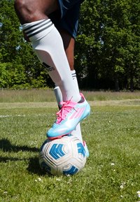 Blue and pink soccer cleat resting on a black and white soccer ball, with white knee-high socks featuring black stripes, on grass.