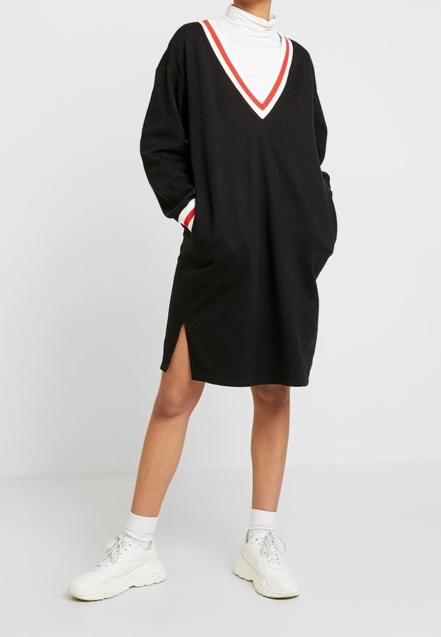 Personne portant une robe noire à manches longues avec un liseré blanc et rouge, un col roulé blanc, des chaussettes blanches et des baskets blanches épaisses.