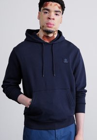 Marineblaue Kapuzenjacke aus weichem Stoff, ausgestattet mit einer Kängurutasche, einer verstellbaren Kapuze und einem kleinen Logopatch auf der Brust.