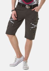 Khaki Cargo-Shorts mit mehreren Reißverschlusstaschen, kontrastierenden Nähten und sichtbaren Logodetails. Getragen mit weißen Sneakers.