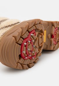 Suela de goma marrón con puntas rojas, que presenta un patrón texturizado para mayor agarre. El material superior es beige con un diseño a rayas.