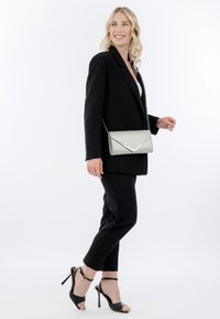 Lachende vrouw in een zwarte blazer en broek, met een zilveren schoudertas, en zwarte sandalen met hoge hakken, lopend tegen een witte achtergrond.