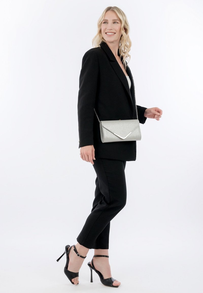 Lachende vrouw in een zwarte blazer en broek, met een zilveren schoudertas, en zwarte sandalen met hoge hakken, lopend tegen een witte achtergrond.