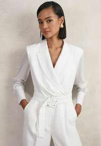 Balts pielāgots kombinezons ar pārklājuma dizainu, garām piedurknēm un jostu viduklī. Raksturo gluds audums un V veida izgriezums. Zelta paziņojuma auskari.