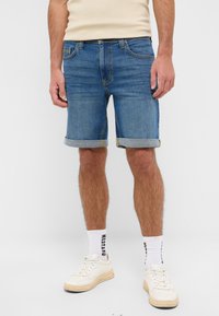 Shorts de mezclilla en azul medio con dobladillos enrollados y costuras en contraste. Combinados con zapatos deportivos blancos y calcetines de longitud media con un logo.