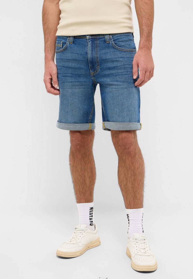 Shorts de mezclilla en azul medio con dobladillos enrollados y costuras en contraste. Combinados con zapatos deportivos blancos y calcetines de longitud media con un logo.