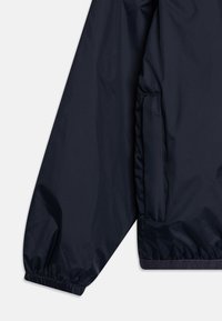 Giacca a vento blu navy con polsini elasticizzati, una tasca laterale e una texture di tessuto morbido e leggero.