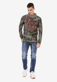 Langarm-Graphic-Sweatshirt mit einem Schädel-Design in Grau, Rot und Grün sowie Text. Kombiniert mit zerrissenen blauen Jeans und weißen Sneakers.