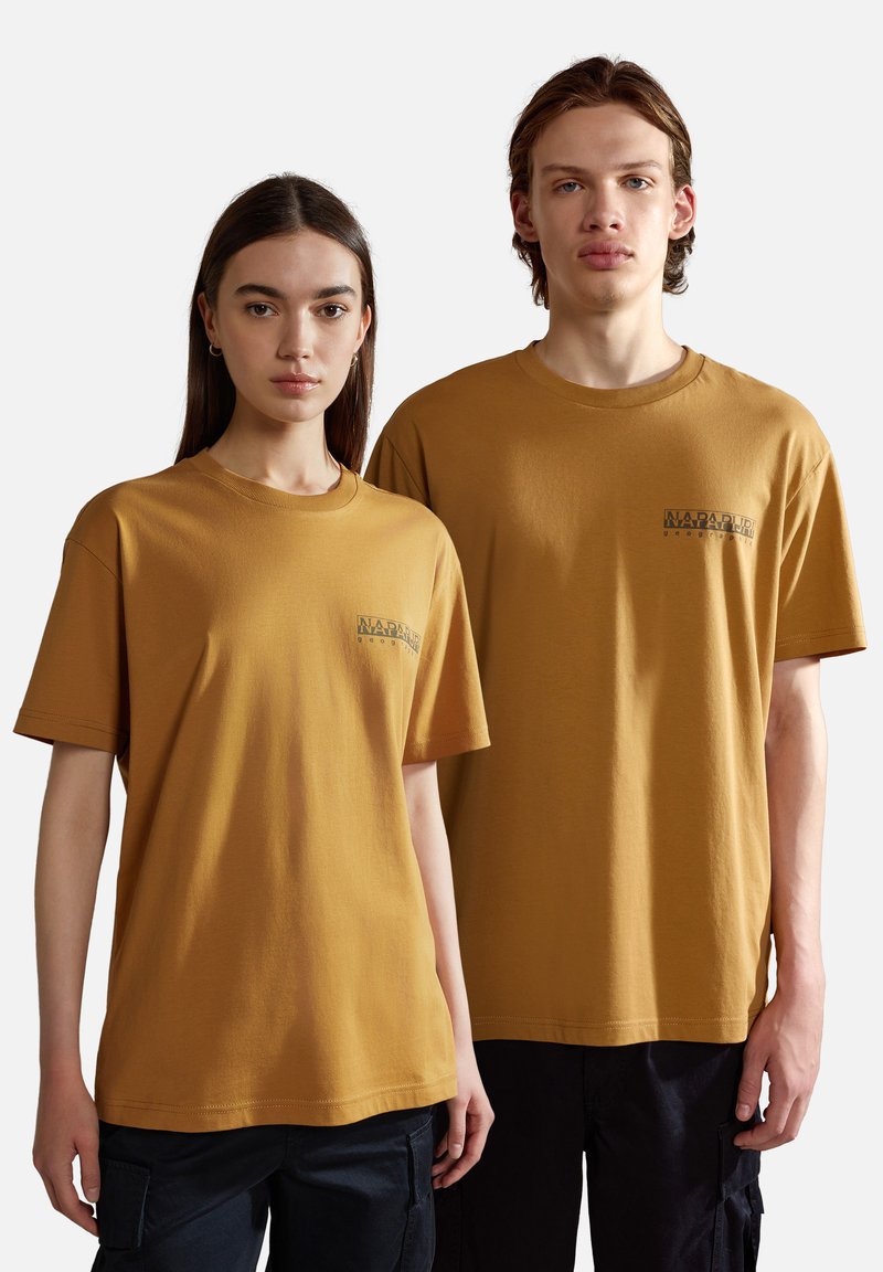 Dos personas llevan camisetas de algodón de color mostaza, de manga corta. Las camisetas cuentan con un pequeño logotipo en la parte izquierda del pecho.
