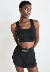 Mujer atlética con un sujetador deportivo negro y pantalones cortos negros de Calvin Klein, de pie con las manos detrás de la espalda contra un fondo blanco liso.