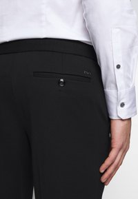 Mies, jolla on mustat juhlahousut, joissa on napillinen takatasku, ja valkoinen, pitkähihainen paitapusero, jonka hihansuut ovat napilliset.