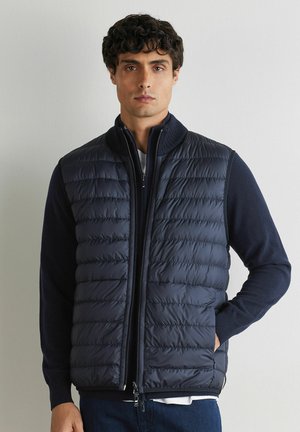 Uomo con capelli corti e ricci che indossa un gilet imbottito blu navy sopra un maglione blu navy, in piedi con una mano nella tasca del gilet contro un muro semplice.