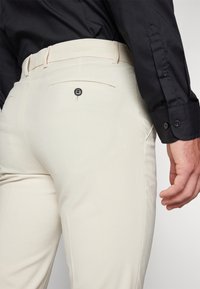 Beige szövetnadrág, hátul zsebbel és fekete gombbal, slim fit kialakítással, egy fekete hosszú ujjú ing mellett bemutatva.
