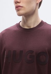 Bordeauxrød sweatshirt med et hævet sort logo punchet med ordet "HUGO." Stoffet virker blødt med en glat tekstur og rund hals.