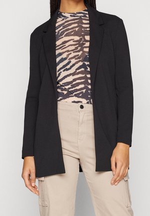 Femme portant un blazer noir ouvert, un top imprimé animalier et un pantalon cargo beige avec poches latérales et fermeture à boutons devant.