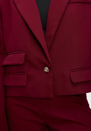 Blazer ajusté bourgogne avec une fermeture à un bouton, deux poches avant et un tissu texturé. Les accents incluent un design de bouton incurvé.