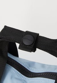 Sangle noire avec un bouton de fermeture circulaire noir sur un sac bleu clair. Le tissu présente une texture lisse avec un détail de surpiqûre contrastant.