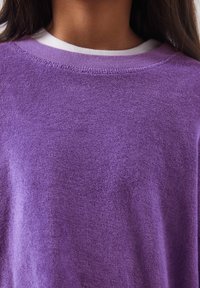 Gros plan sur une personne portant un pull violet texturé par-dessus un t-shirt blanc à col rond, mettant en avant l'encolure et le haut de la poitrine.