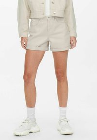 Shorts en denim beige avec des ourlets retroussés, dotés de deux poches avant et d'une fermeture à bouton, associés à des baskets blanches et des chaussettes.