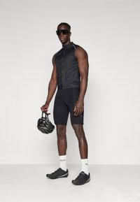 Gilet de cyclisme sans manches noir sur un short rembourré noir, tenant un casque de vélo noir. Porté avec des chaussettes blanches et des chaussures de sport noires.