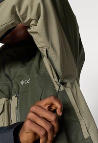 Vandeniui atspari striukė su žaliais ir chaki spalvos paneliais, turinti užtrauktuką kišenėje ir raišteliu detale rankogalyje, leidžiančia reguliuoti dydį.