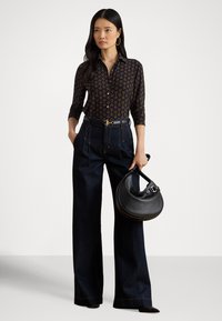 Sieviete, kas valkā rakstainu melnu blūzi, tumšas krāsas augsta vidukļa platās džinsa bikses, tur melnu pusmēness formas rokassomu, stāv pret vienkāršu fona sienu.