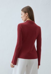 Hosszú ujjú, mélyvörös bordázott anyagból készült ing magas nyakkal és testhezálló fazonnal, fehér nadrággal párosítva. Sima textúra, minimalista részletezés.