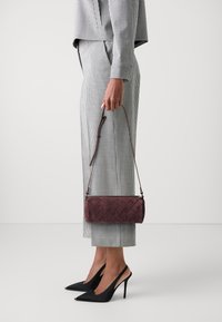 Borsa intrecciata di un viola scuro con una tracolla removibile, tenuta in una mano, abbinata a un completo grigio a scacchi e tacchi a spillo neri.