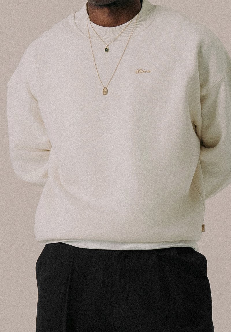 Personne portant un sweat-shirt crème avec un logo doré "Blkots" et deux colliers en or, associé à un pantalon noir sur un fond uni.