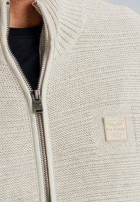 Lichtcrème gebreide jas met een textuurpatroon, voorzien van een hoge kraag, volledige rits en een vierkante logo patch.