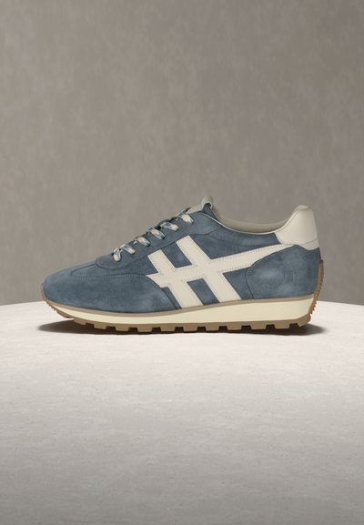 Μπλε σουέντ αθλητικό παπούτσι με λευκές πλευρικές ρίγες, μπεζ φτέρνα και σόλα από καουτσούκ σε χρώμα καραμέλας, πάνω σε ανοιχτόχρωμη επιφάνεια με ουδέτερο γκρι φόντο.