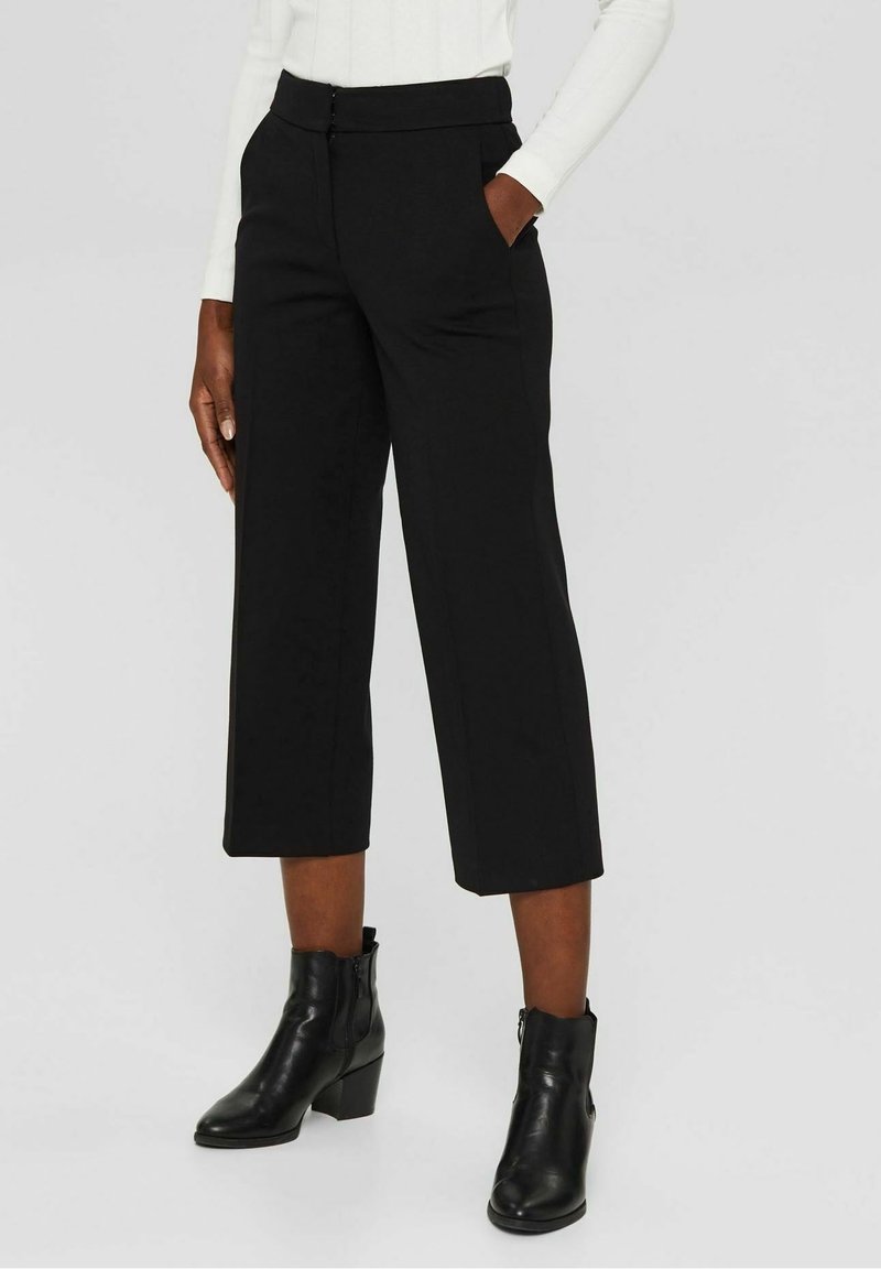 Pantalon noir court avec une coupe ajustée, taille plate, poches latérales et tissu lisse. Porté avec des bottines noires.