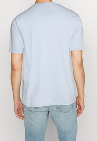 Mann trägt ein hellblaues T-Shirt mit kurzen Ärmeln und ausgewaschene blaue Jeans, von hinten vor einem einfarbigen hellen Hintergrund gezeigt.