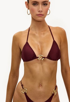 Femme portant un bikini bordeaux avec des détails métalliques en spirale dorée sur le haut et le bas, ainsi que des boucles d'oreilles créoles dorées, se tenant devant un fond blanc.