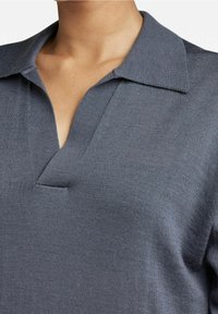 Grå stickad polo med krage och en V-formad öppning fram. Tyget har en slät, strukturerad yta.