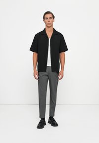 Camicetta nera a maniche corte con bottoni sopra una canotta bianca, abbinata a pantaloni grigi e scarpe nere. Tessuto liscio, vestibilità slim, design minimale.