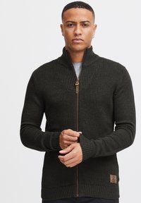 Suéter de punto de color verde oscuro con cremallera, con cuello y puños de canalé, que presenta un cuerpo texturizado y un parche de logo de cuero marrón claro en el dobladillo.