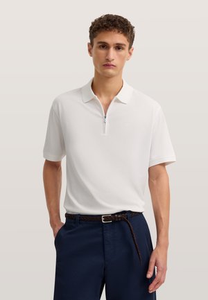 Junger Mann mit kurzen lockigen Haaren, trägt ein weißes kurzärmeliges Polohemd mit Reißverschluss und marineblaue Hose mit geflochtenem Gürtel.