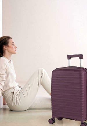 Maleta rígida morada con líneas onduladas texturizadas, dos ruedas lisas y un asa telescópica, colocada junto a una figura sentada.