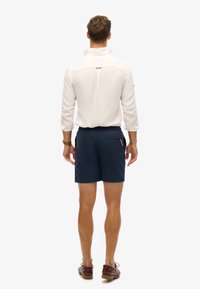 Camisa blanca de lino con cuello alto y abertura trasera, combinada con pantalones cortos azul marino con un bolsillo trasero y cintura elástica. Calzado casual.
